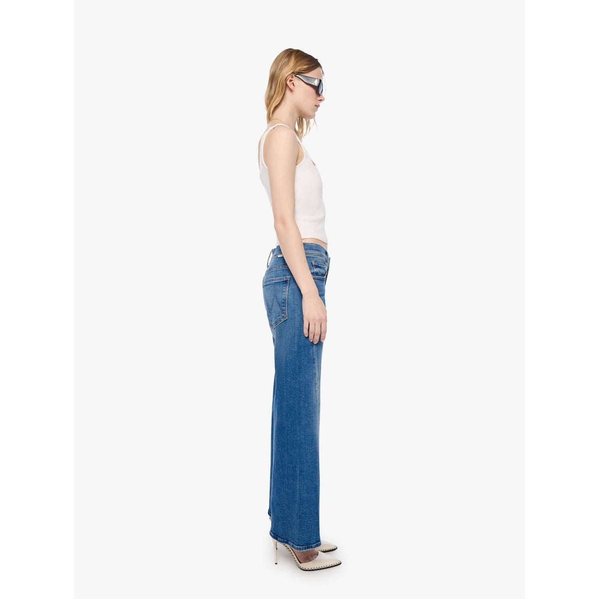 A woman shown in profile wearing blue wide-leg jeans, a white ribbed tank top, and pointed shoes against a plain light gray studio background.