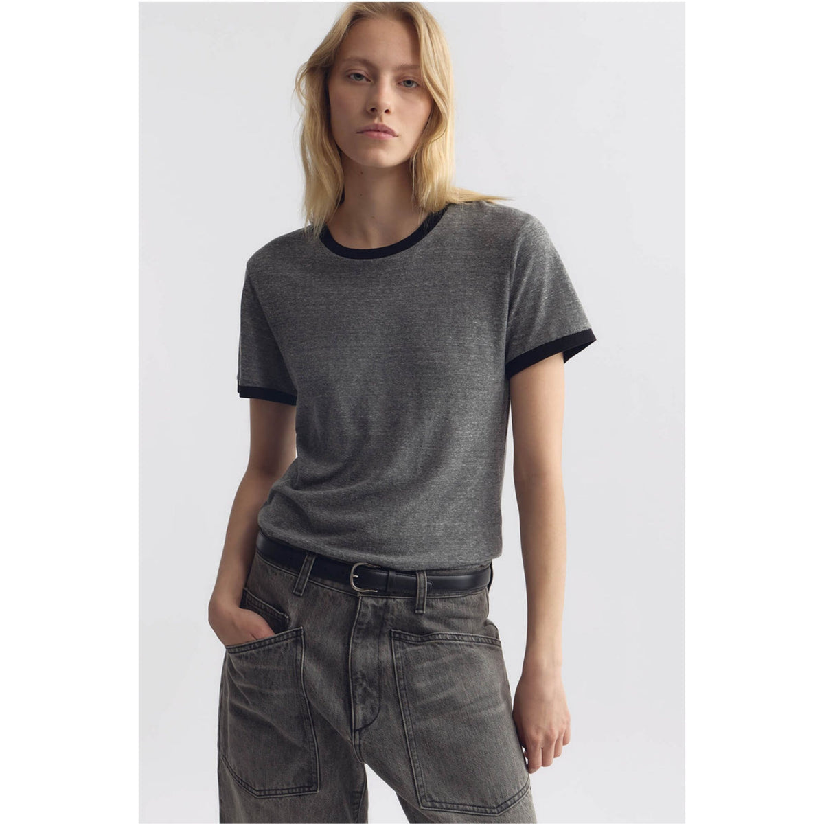 A woman modeling a dark gray ringer t-shirt with black trim on the crew neck and sleeve cuffs, paired with gray jeans and a black belt, against a light gray studio background.