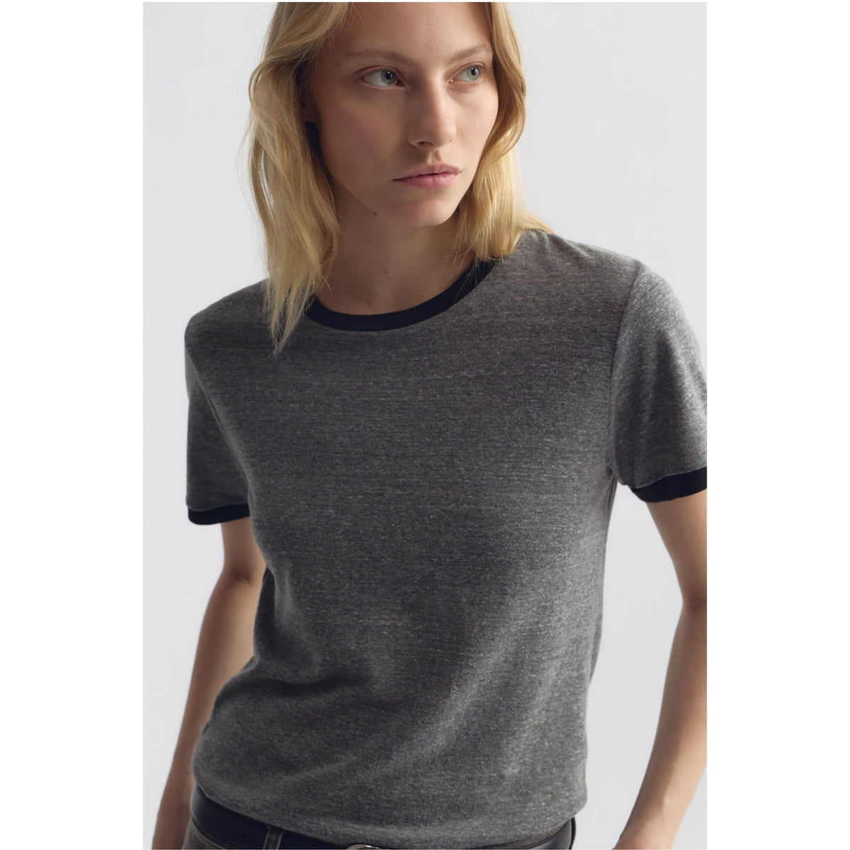 A woman wearing a dark heather gray short-sleeve ringer T-shirt with black ribbed trim at the crew neck and sleeve cuffs, photographed against a light gray studio background.