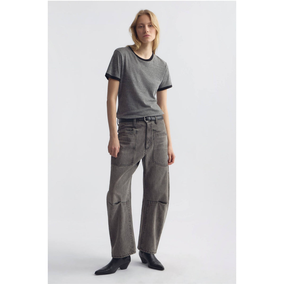 A model stands against a light gray studio background wearing a gray ringer T-shirt with black trim, wide-leg gray jeans with patch pockets, and black ankle boots.