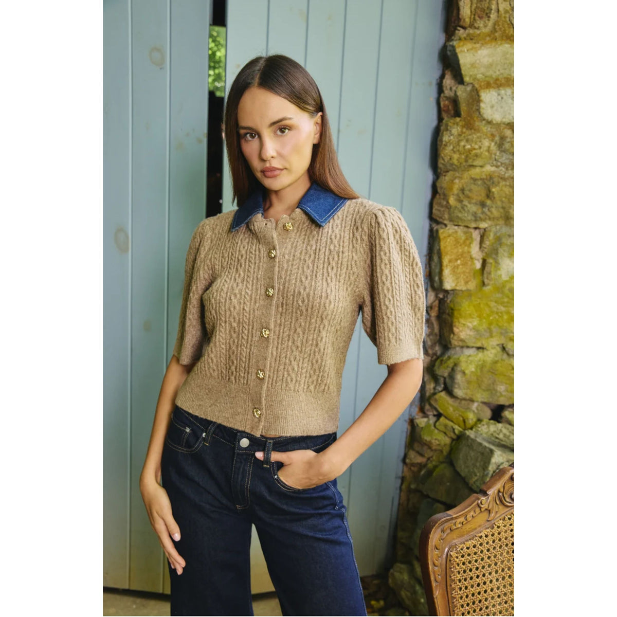A woman modeling a short-sleeve cable-knit cardigan with a blue collar and gold buttons, paired with dark blue jeans, standing in front of a light blue wooden door and a stone wall.