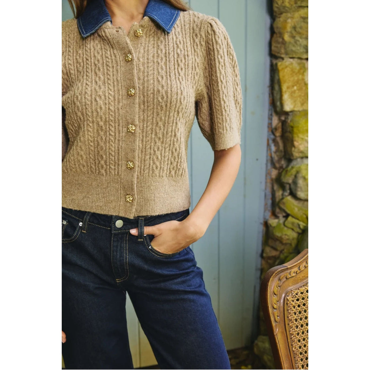 A woman wearing a beige cable-knit short-sleeve cardigan with a blue denim collar and gold buttons, paired with dark blue jeans, standing in front of a rustic stone wall and blue paneled door.