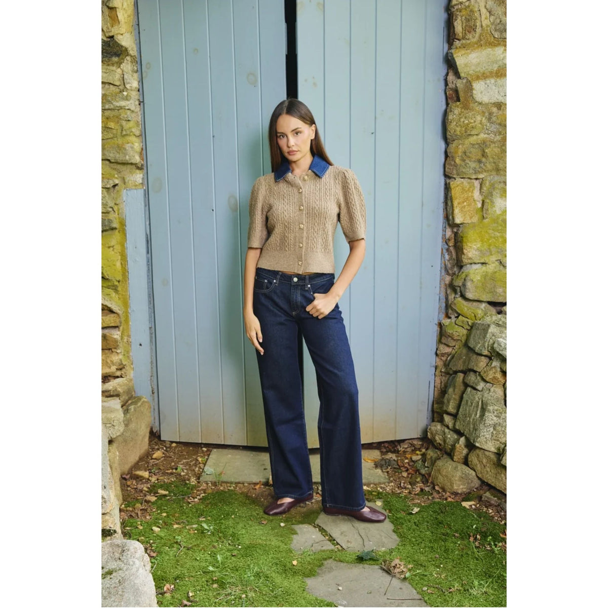 A woman stands outdoors in front of a light blue wooden door, wearing a beige short-sleeve knit polo shirt with a blue collar and dark navy wide-leg jeans.