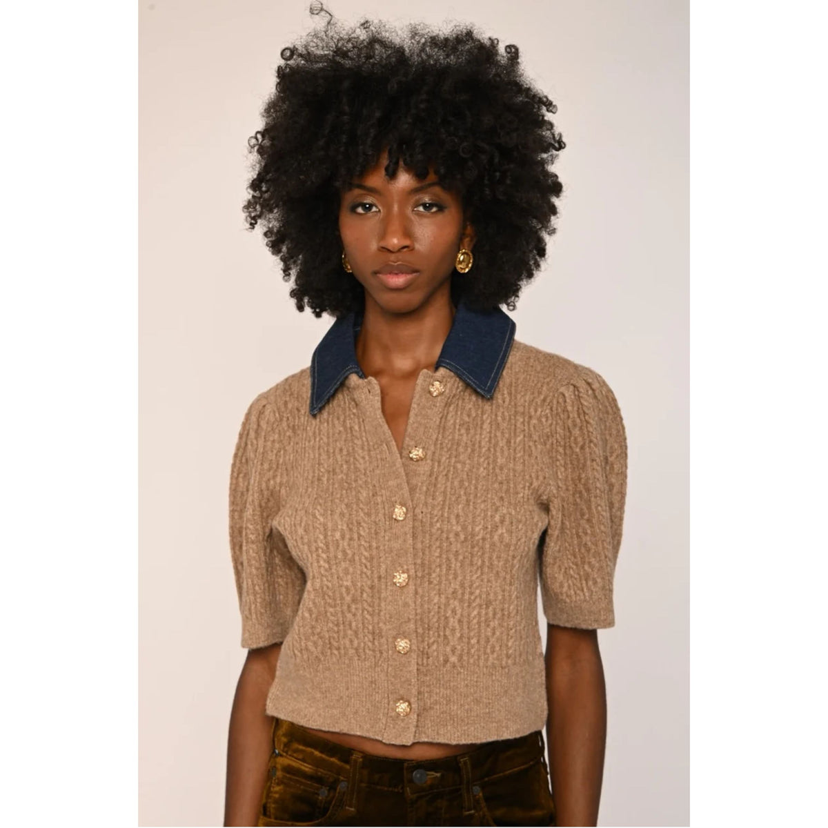 A model with a large black afro wearing a beige cable-knit cropped short-sleeve top with a contrasting navy blue collar and gold buttons, paired with brown pants, against a plain light background.