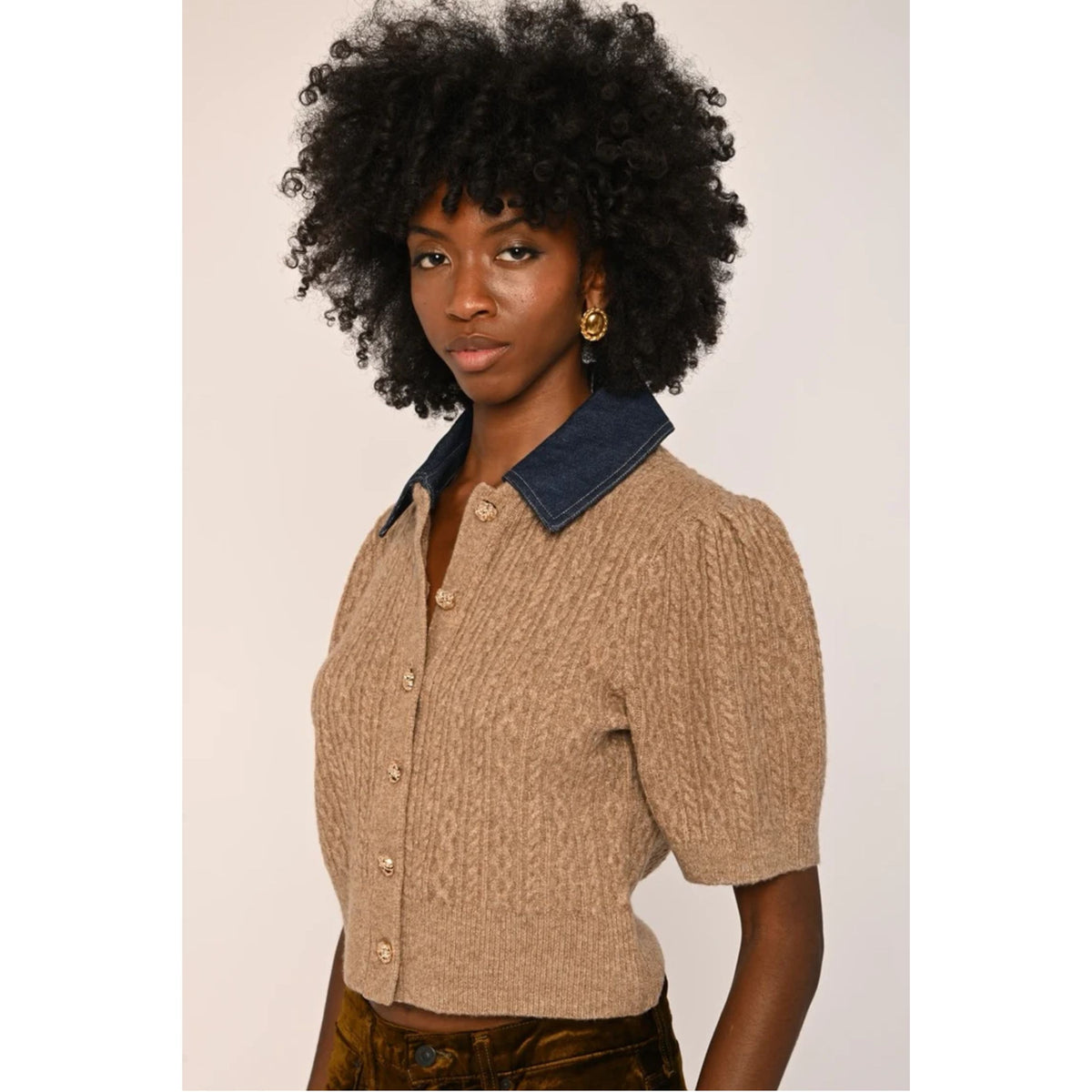 A model with a voluminous black afro hairstyle wearing a beige textured short-sleeve button-up cardigan with a dark navy blue collar, paired with brown patterned trousers, against a plain light beige background.