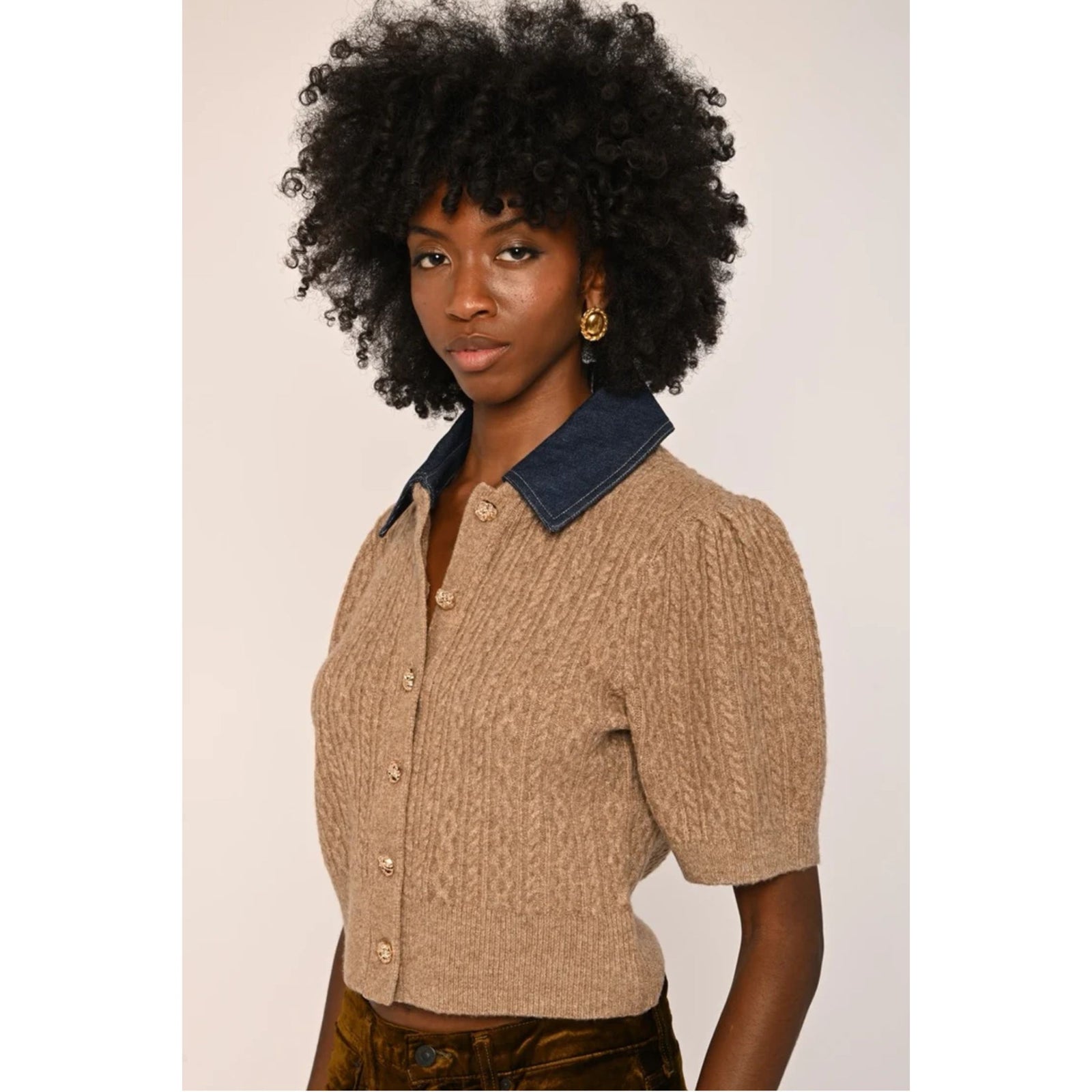 A model with a large black afro wearing a beige cable-knit cropped short-sleeve top with a contrasting navy blue collar and gold buttons, paired with brown pants, against a plain light background.