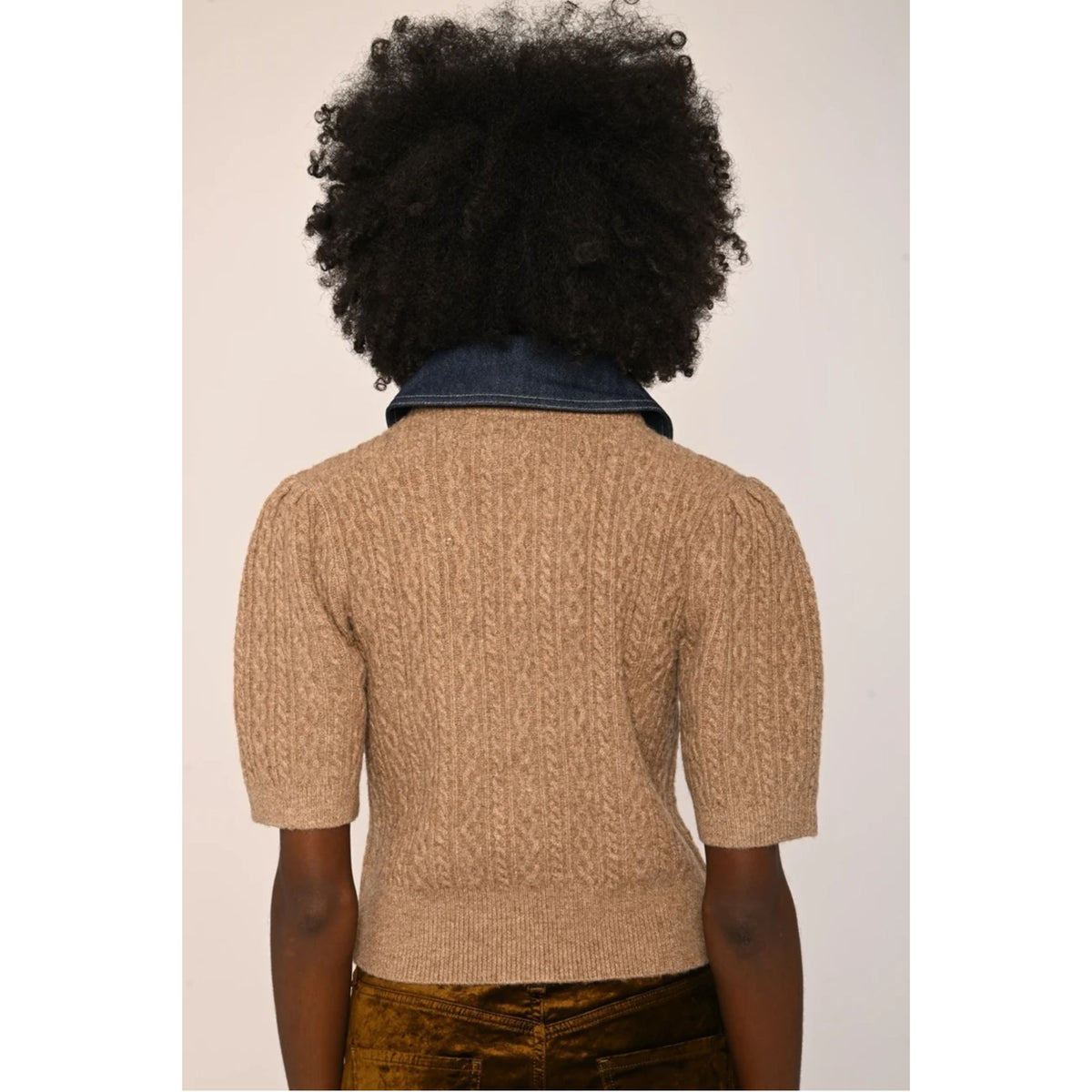 A person shown from the back wearing a beige short-sleeve textured knit sweater over a dark blue denim collar, paired with brown pants, against a plain light background.
