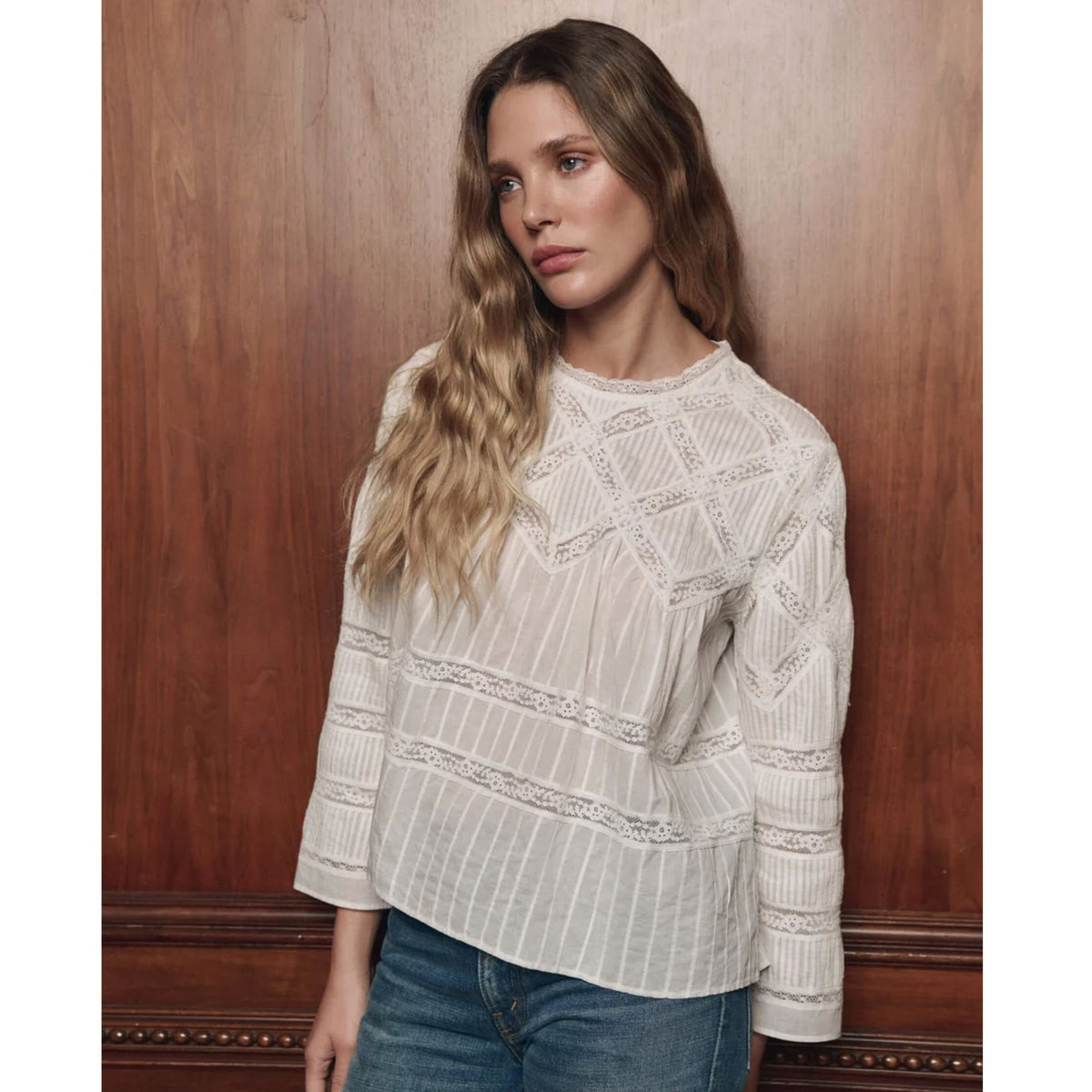 A woman with long wavy light brown hair wearing a cream-colored textured knit sweater with geometric patterns and blue jeans, standing against a dark wood-paneled wall.