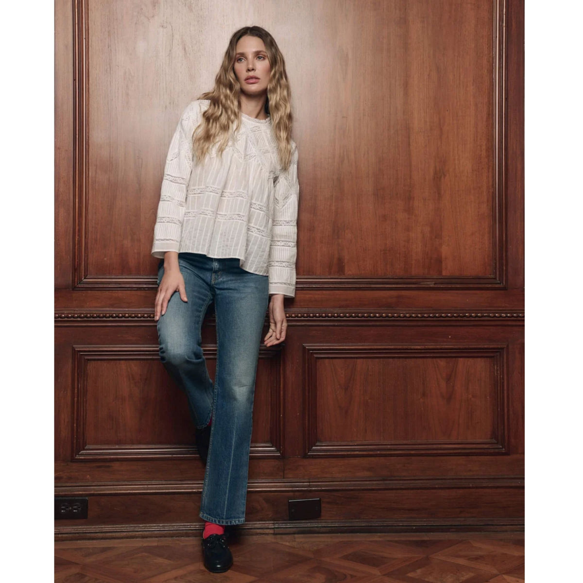 A woman stands against a dark wood paneled wall wearing a cream textured long-sleeve top, blue jeans, and black shoes with red socks.
