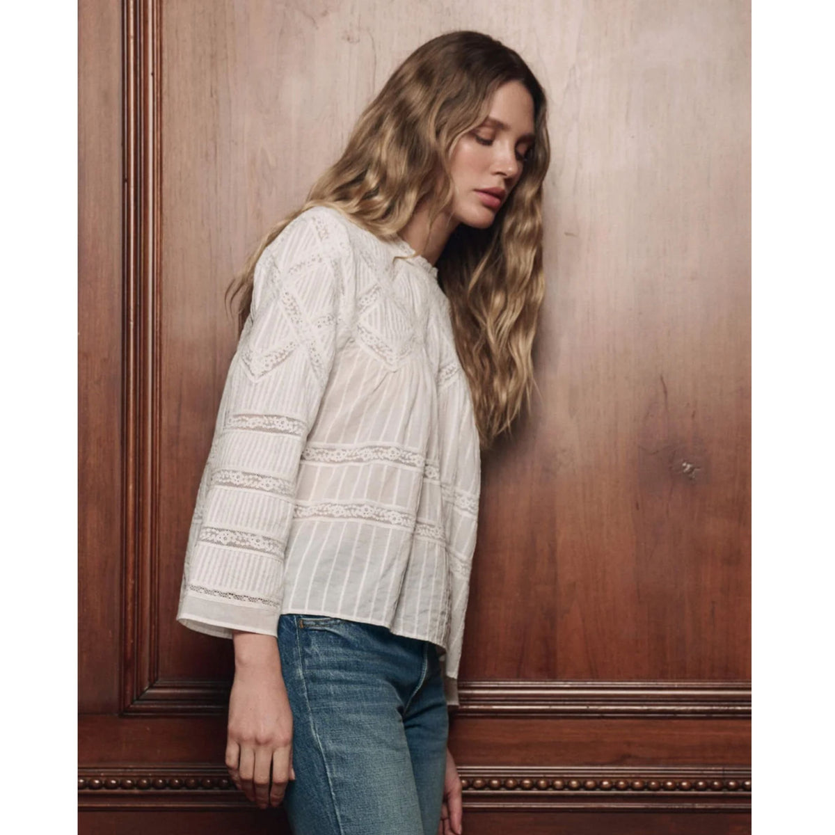 A woman with long wavy blonde hair leans against a warm brown wooden paneled wall, wearing a cream-colored textured knit blouse with lace details and blue jeans.