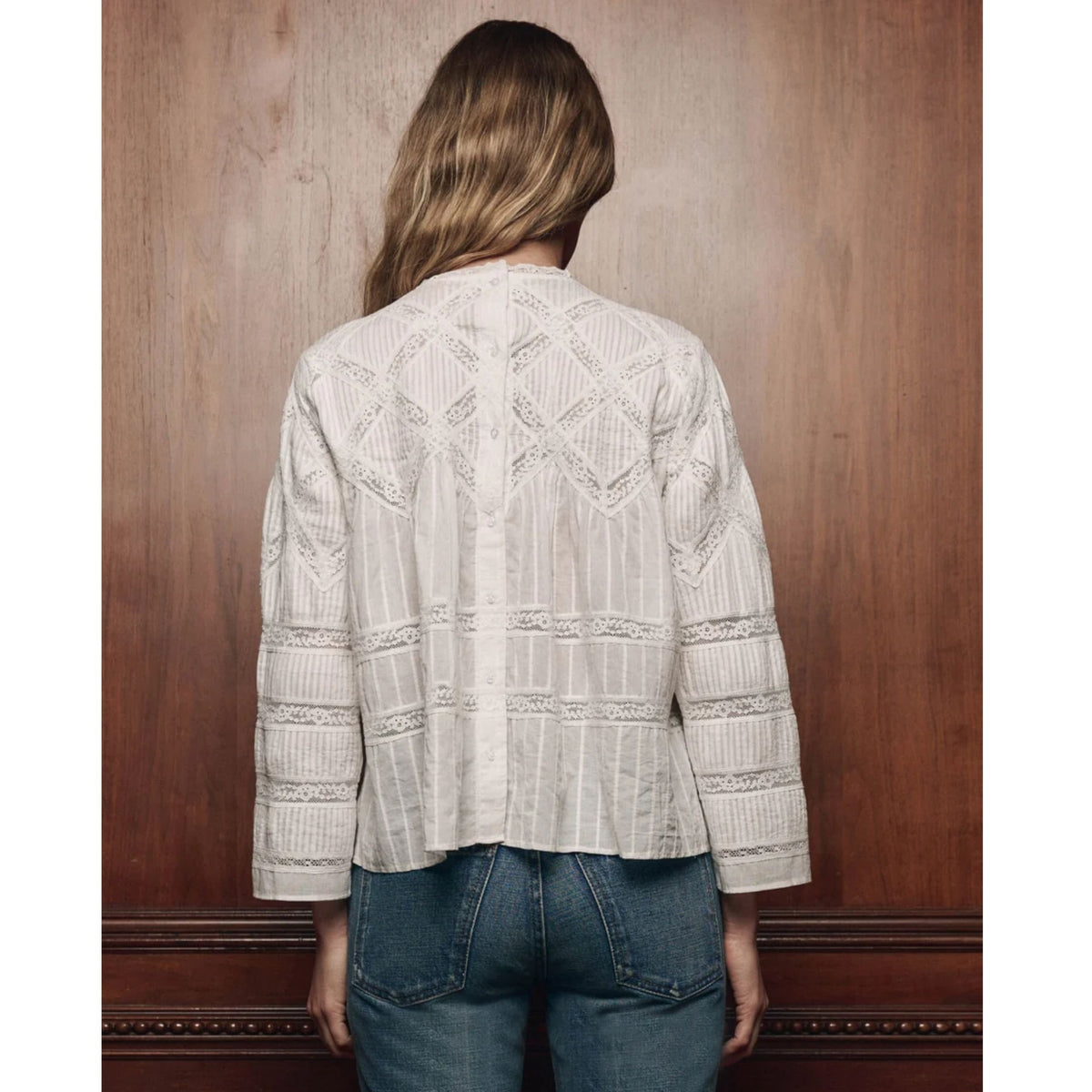 A woman viewed from behind wearing a cream-colored, long-sleeved lace blouse and blue jeans, standing against a dark brown wooden wall.