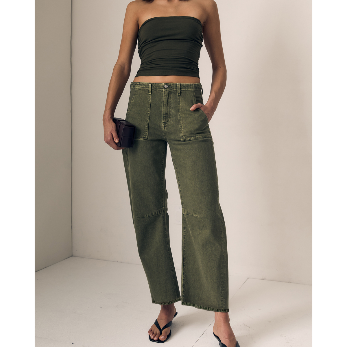 A model wearing olive green wide-leg cropped jeans, a matching olive green strapless crop top, and black sandals, holding a small black clutch against a neutral off-white studio background.