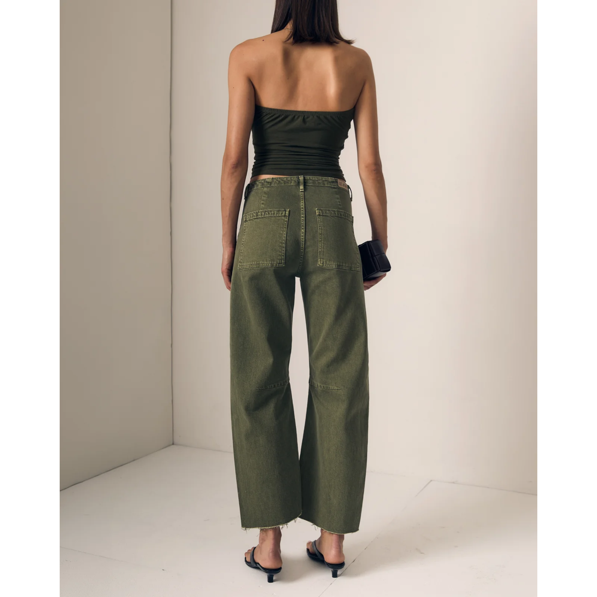 A woman shown from the back wearing olive green wide-leg cropped jeans, a dark olive strapless top, and black heeled sandals in a minimalist beige studio setting.