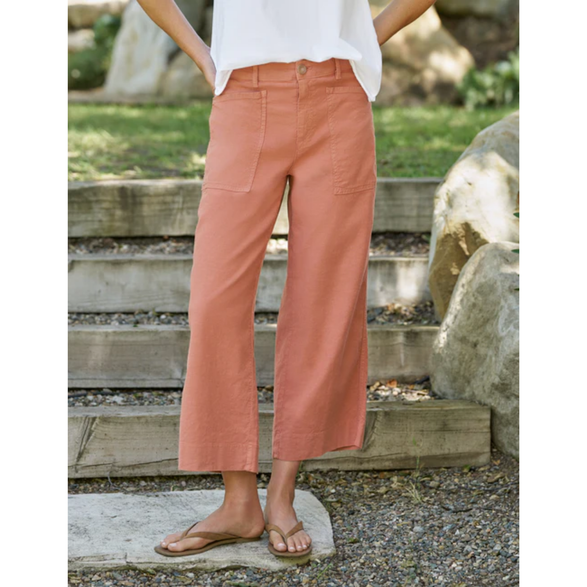 A person wearing wide-leg cropped pants in a muted peach or dusty rose color, paired with a white top and tan flip-flops, standing outdoors on stone steps with rocks and greenery in the background.