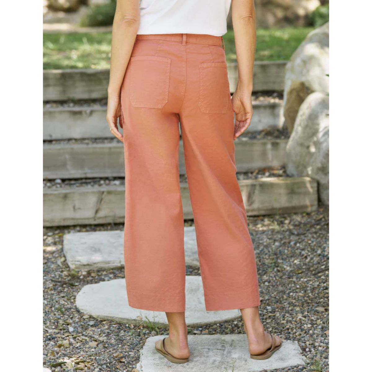 A person shown from the back wearing wide-leg cropped pants in a muted peach or terracotta color, standing outdoors on stone steps with gravel and greenery in the background.