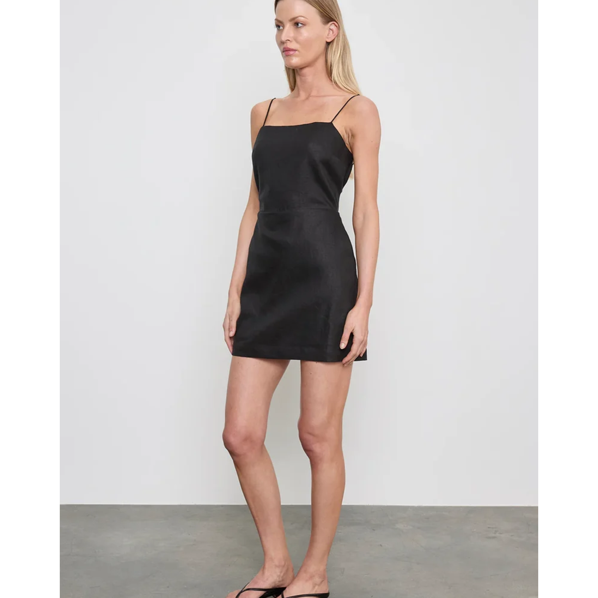 A woman modeling a black spaghetti-strap mini dress against a light gray studio background.