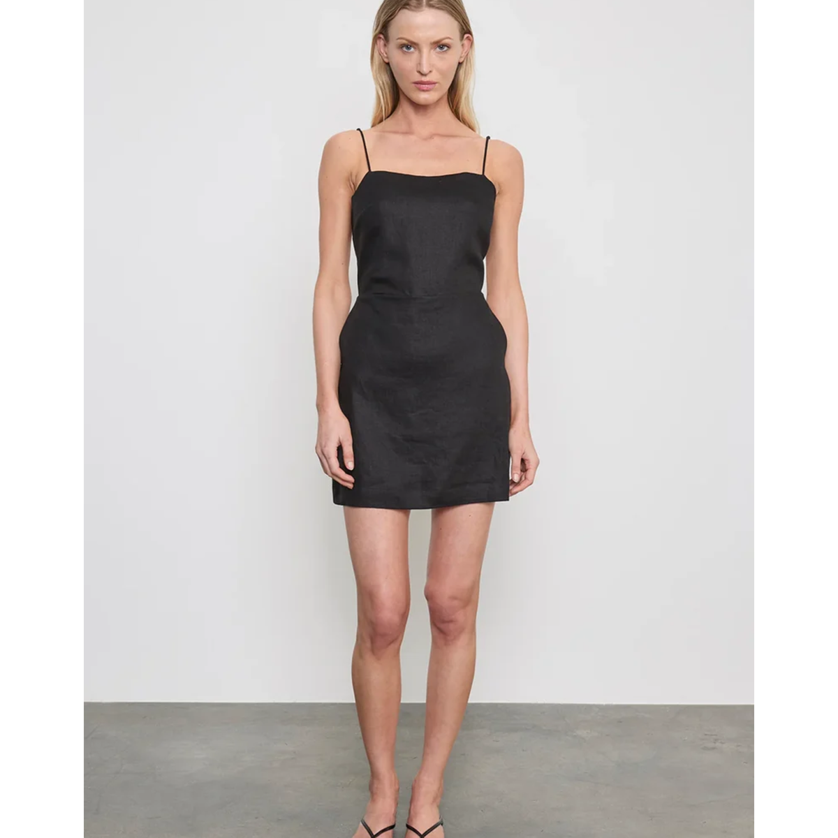 A woman modeling a black spaghetti-strap mini dress against a light gray studio wall and concrete floor.
