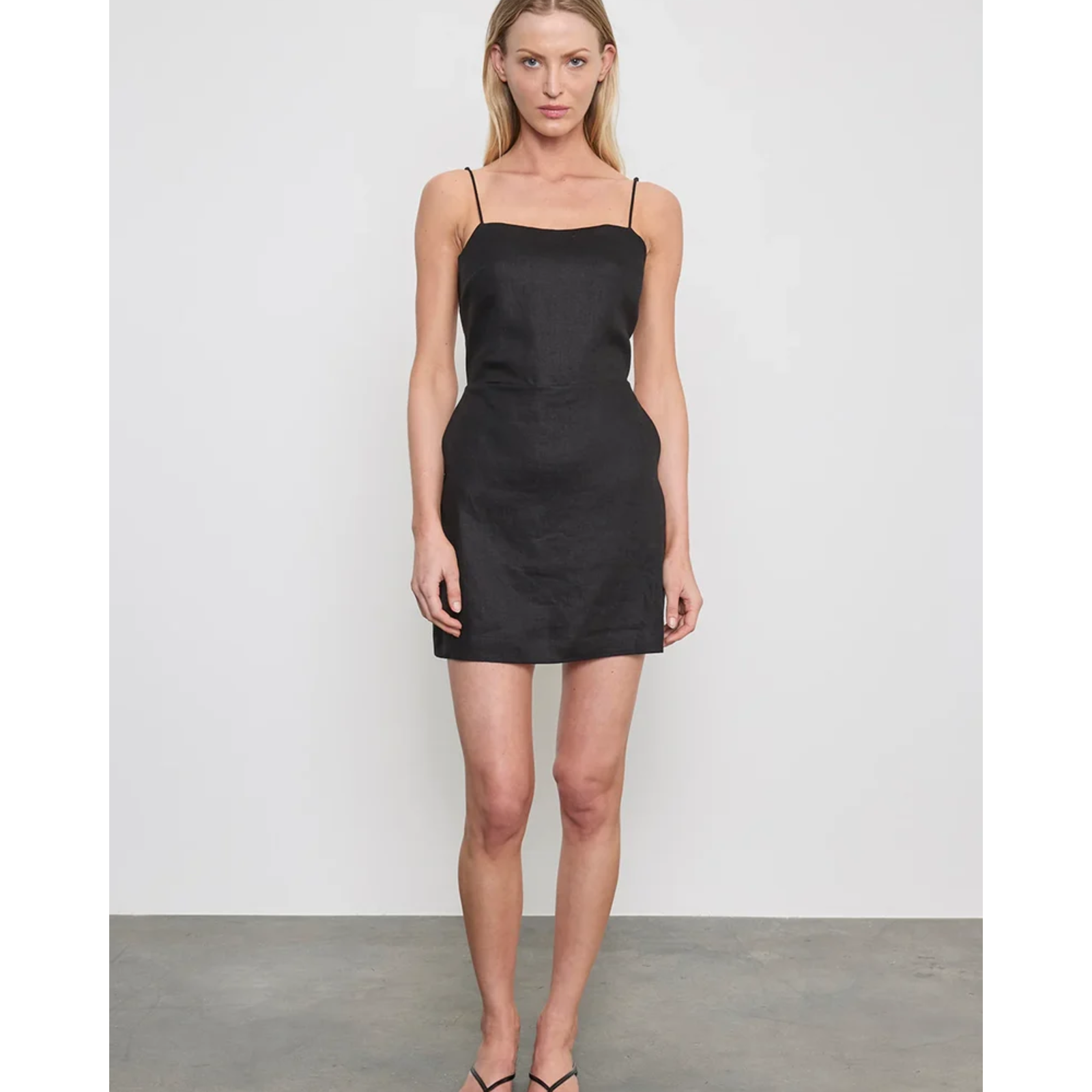 A woman modeling a black spaghetti-strap mini dress against a light gray studio wall and concrete floor.