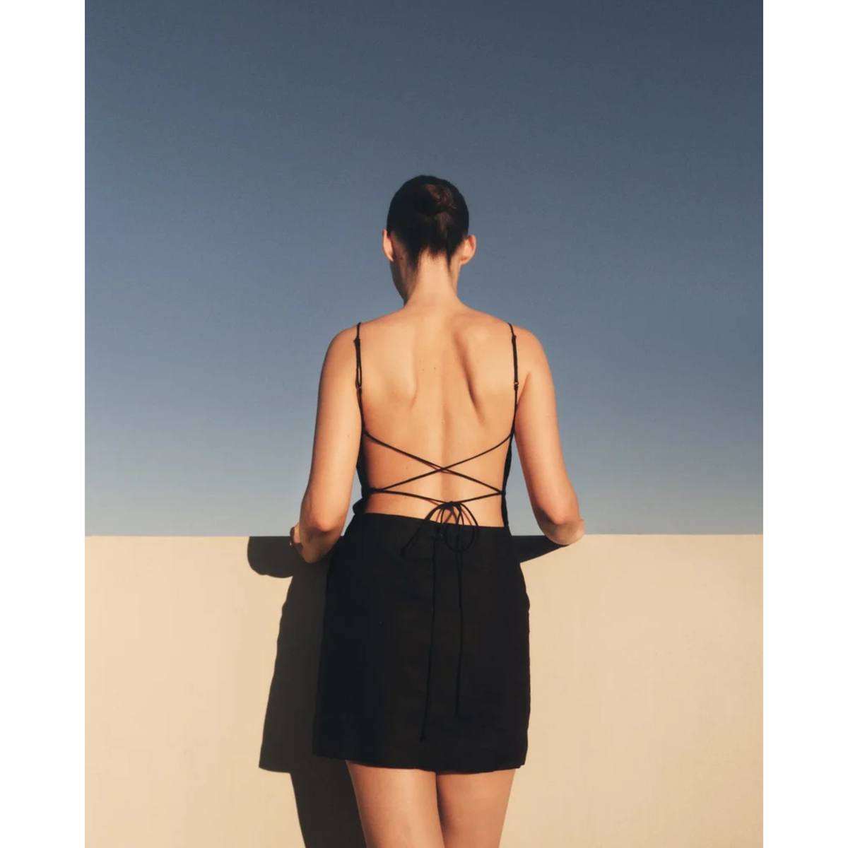 A woman viewed from behind wearing a black backless mini dress with thin shoulder straps and crisscross tie details at the back, standing against a clear blue sky and beige wall.