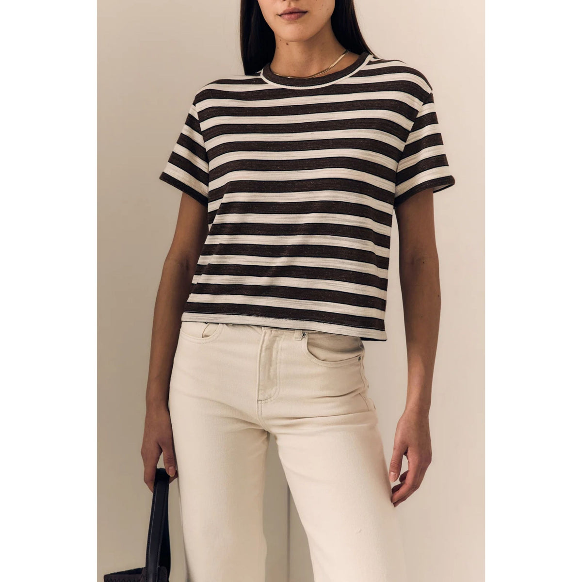 A woman wearing a black and off-white horizontally striped short-sleeve crop top and cream-colored pants, standing against a plain beige wall.