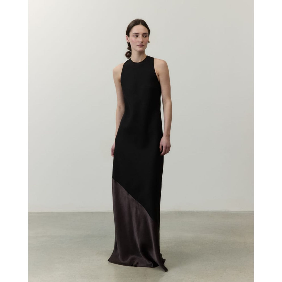 A woman stands in a studio wearing a sleeveless, high-neck floor-length dress. The dress is black with a contrasting dark brown satin lower panel, photographed against a plain light gray wall and concrete floor.