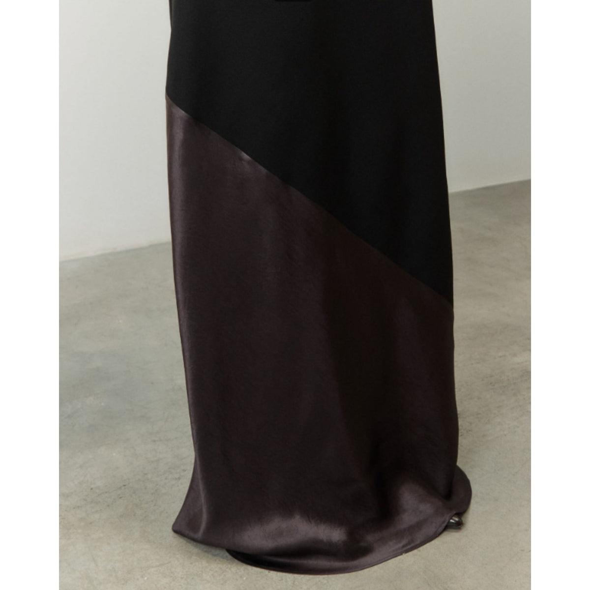 A close-up of a long draped garment or fabric in a color-blocked design, featuring a solid black upper section and a dark brown satin lower section, shown against a neutral studio background.