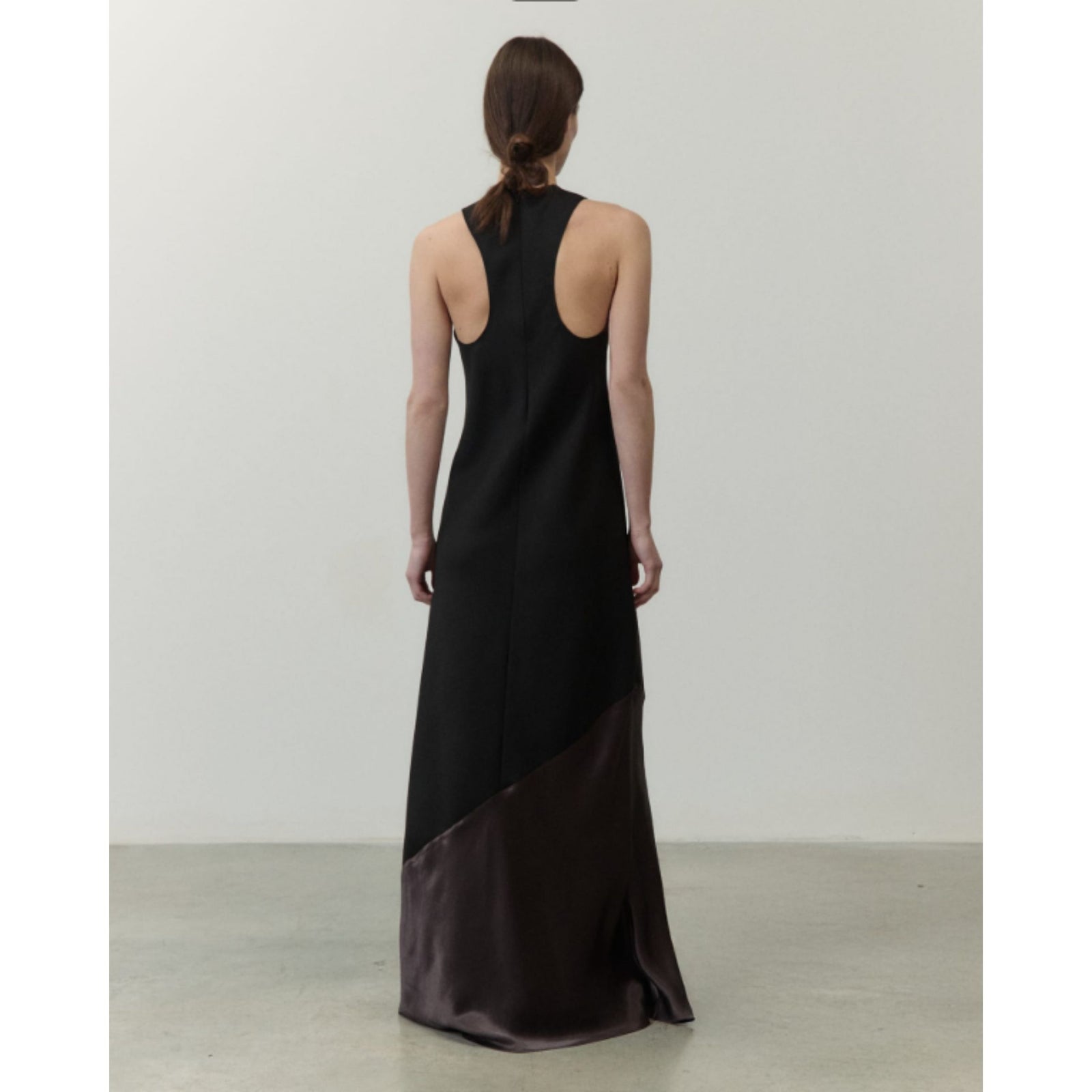 A woman stands in a studio wearing a sleeveless, high-neck floor-length dress. The dress is black with a contrasting dark brown satin lower panel, photographed against a plain light gray wall and concrete floor.