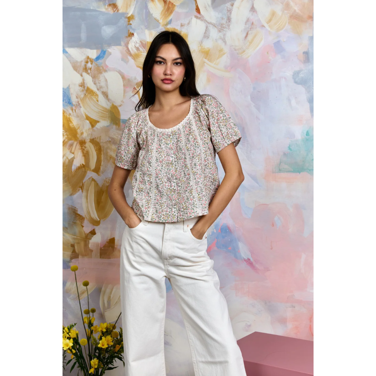 Woman wearing a patterned top and white pants standing against a colorful floral background