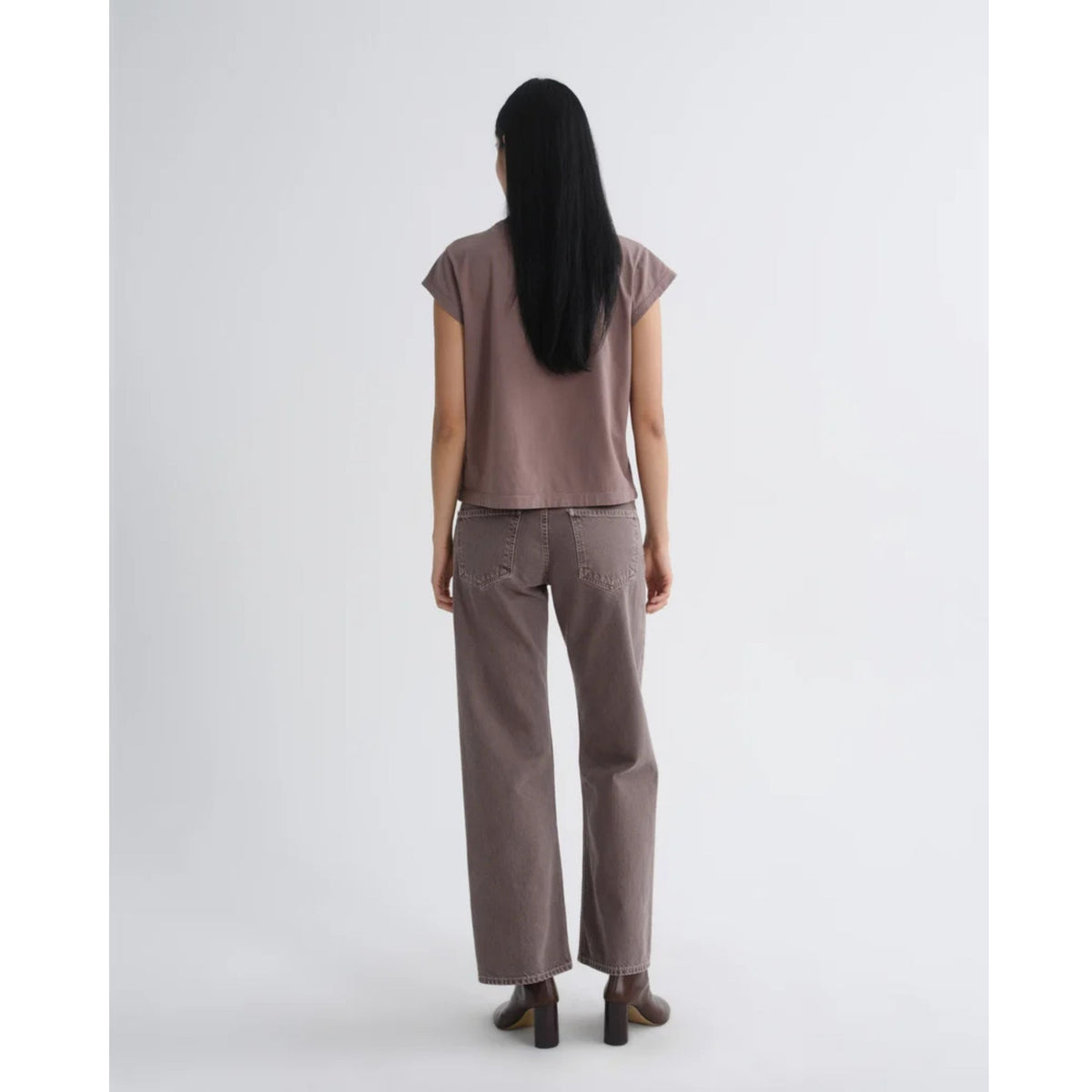 Back view of a model with long dark hair wearing a short-sleeve dusty mauve top and AGOLDE Low Rise Loose jeans with dark boots against a light gray background.