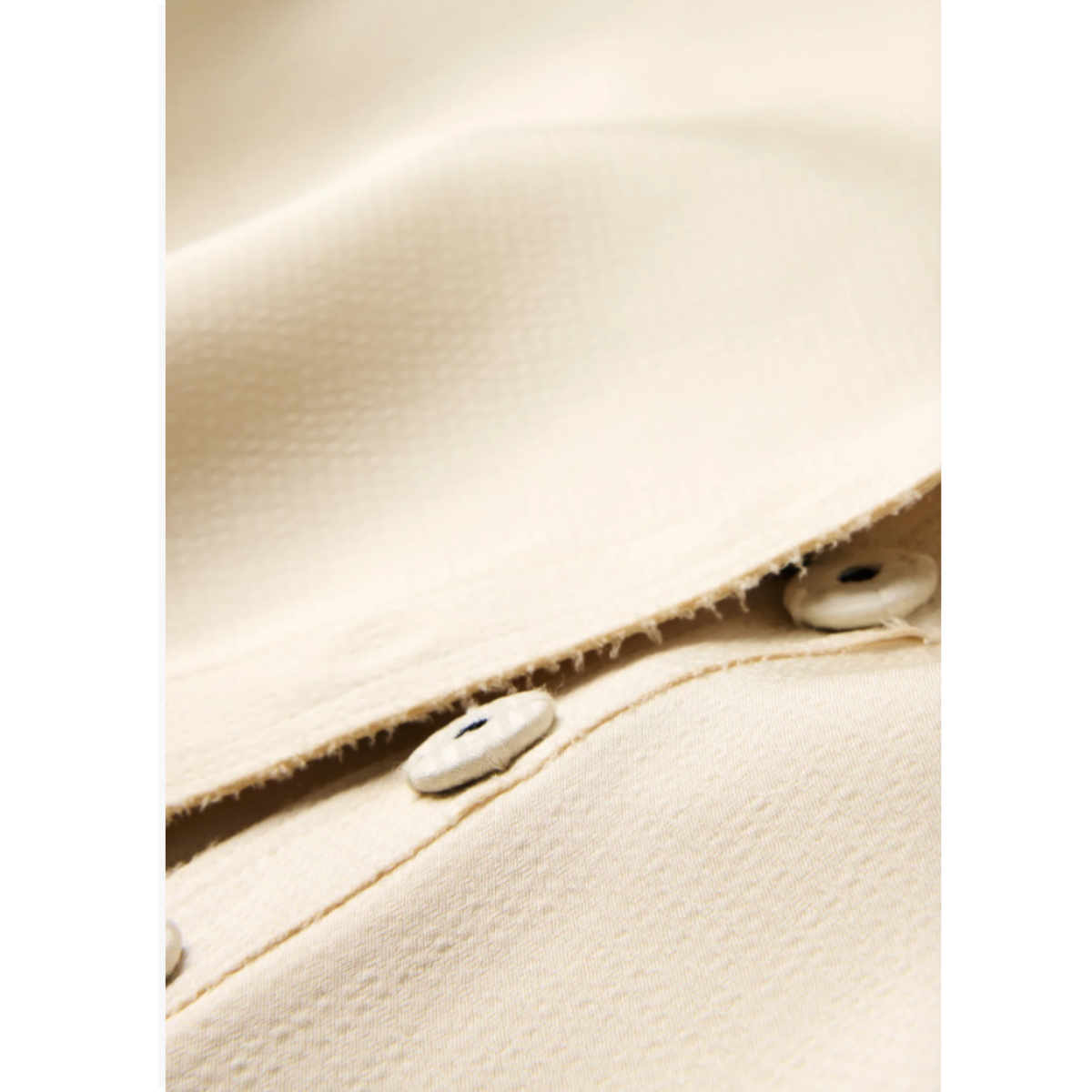Close-up of a cream-colored textured fabric garment with white buttons and a frayed edge.