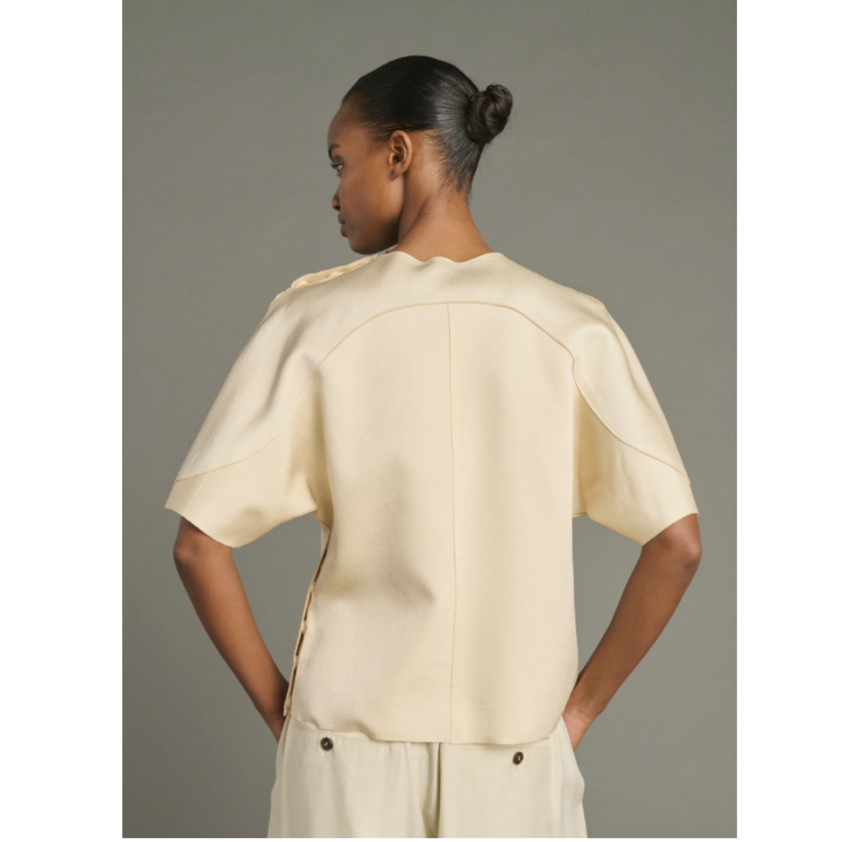 A model shown from the back wearing a cream-colored short-sleeve top and matching pants against a solid gray studio background.