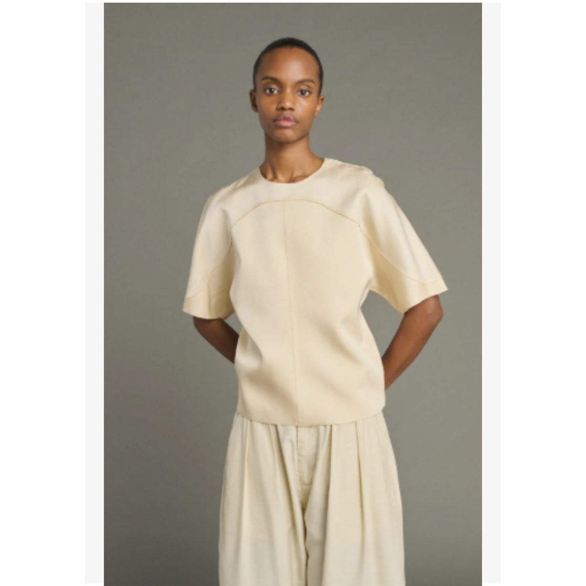 A model wearing a cream-colored short-sleeve top and matching wide-leg trousers against a solid gray studio background.