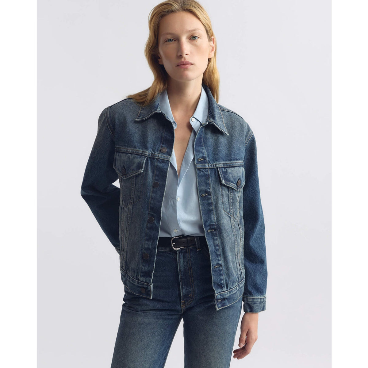 A woman modeling a dark blue denim jacket over a light blue button-up shirt and dark blue jeans, photographed against a plain light gray studio background.