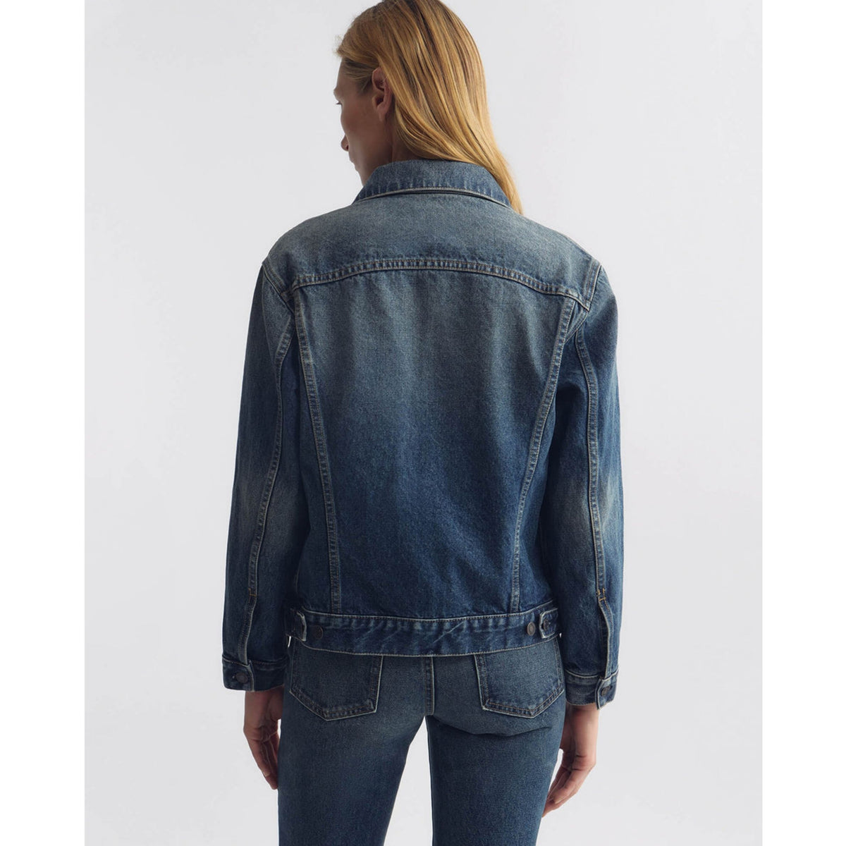 A woman viewed from the back wearing a dark blue denim jacket and matching blue jeans against a plain light gray studio background.