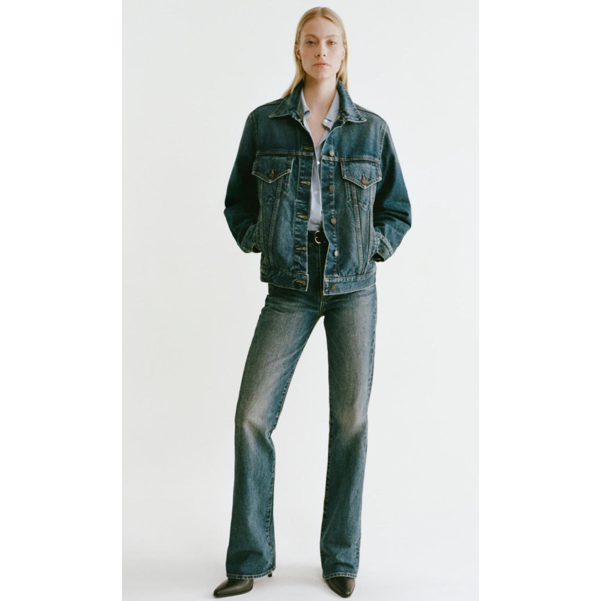 A model stands against a plain light gray studio background wearing a dark blue denim jacket and matching dark blue denim jeans with a white shirt and black shoes.