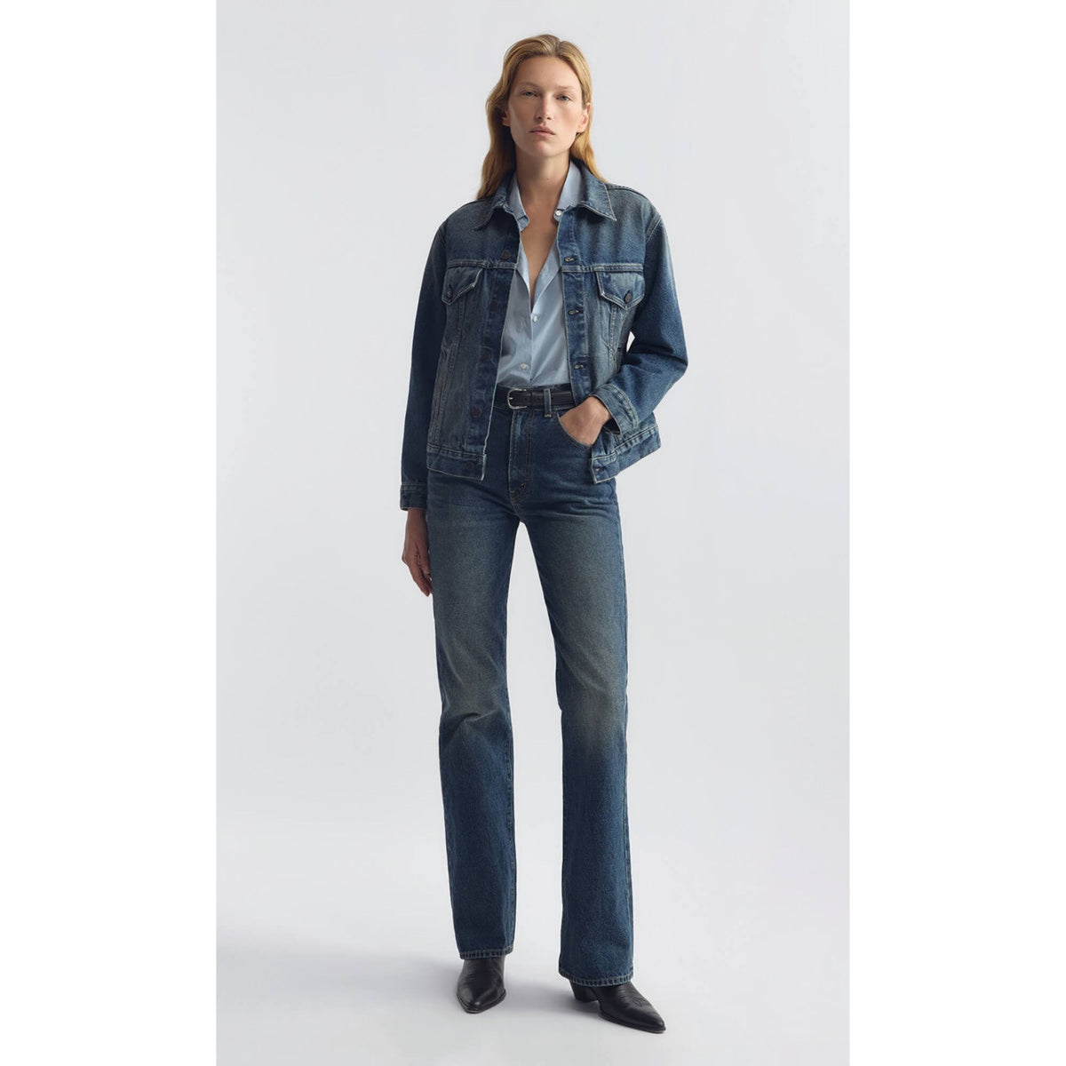 A model stands against a light gray studio background wearing a dark blue denim jacket, matching dark blue jeans, a light blue button-up shirt, and black pointed shoes.