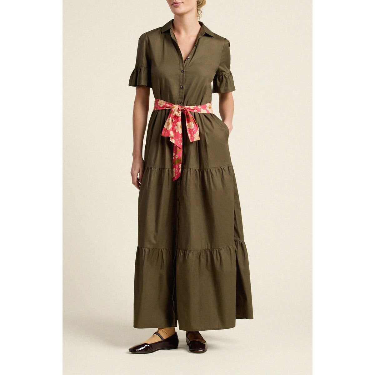 A woman models a dark olive green tiered maxi shirt dress with ruffled short sleeves and a pink floral patterned self-tie belt, paired with dark brown flat shoes against a light beige background.