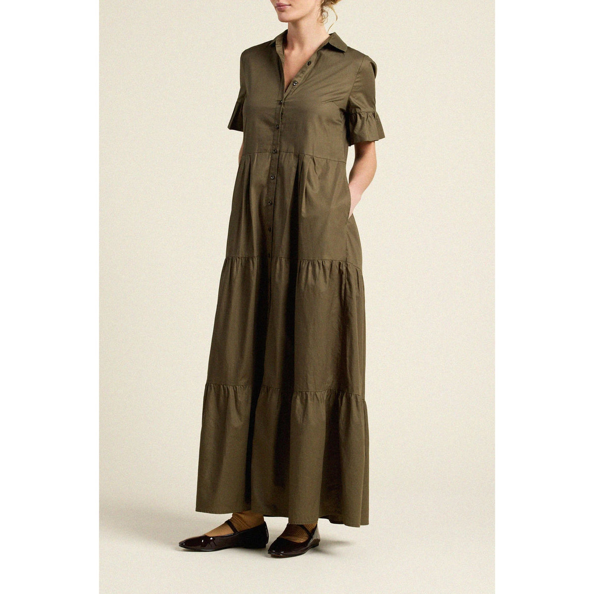 A woman models a short-sleeved, tiered maxi dress in olive green, paired with dark brown shoes, against a plain beige studio background.