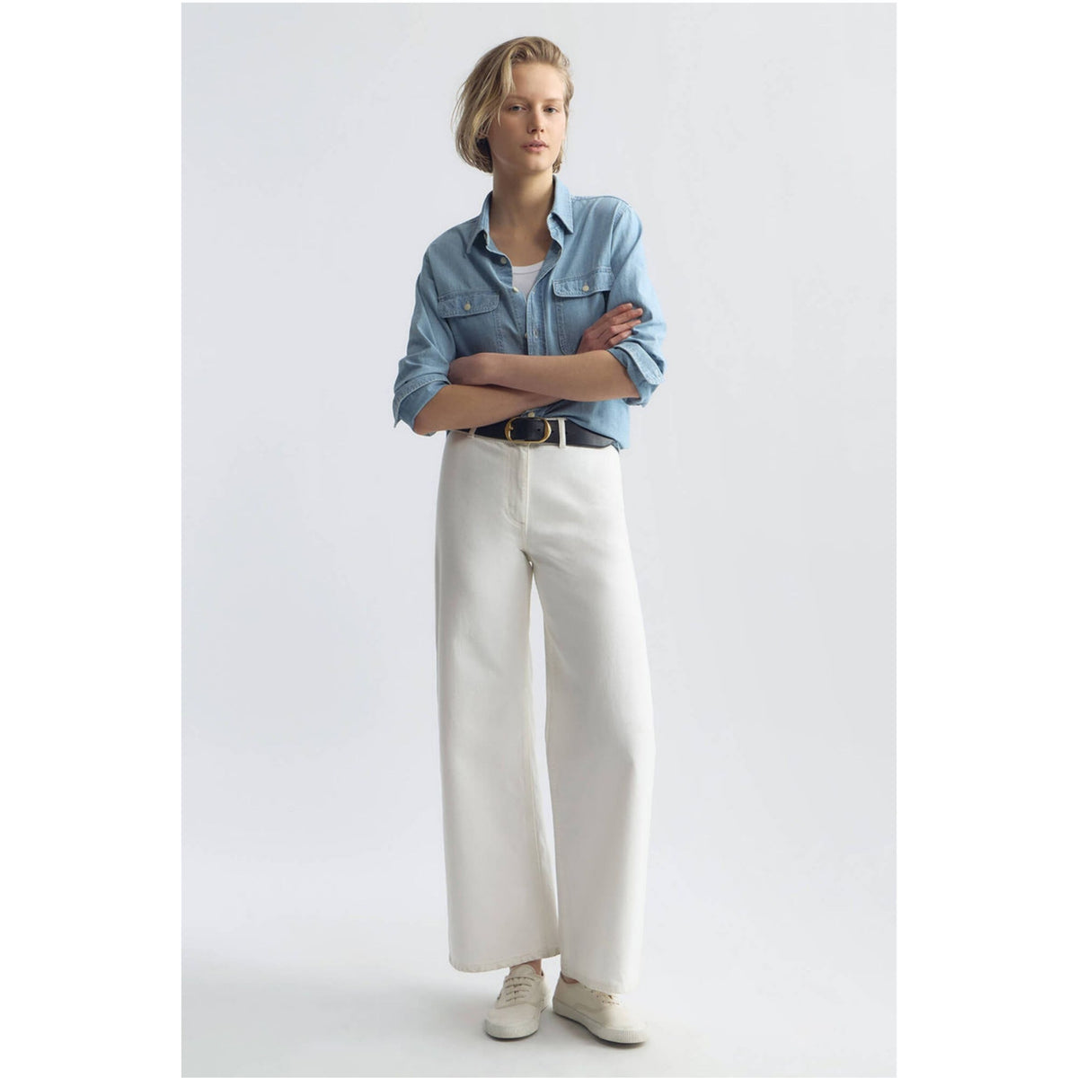 A model stands against a light gray studio background wearing a light blue denim shirt, white wide-leg trousers, a black belt, and white sneakers.