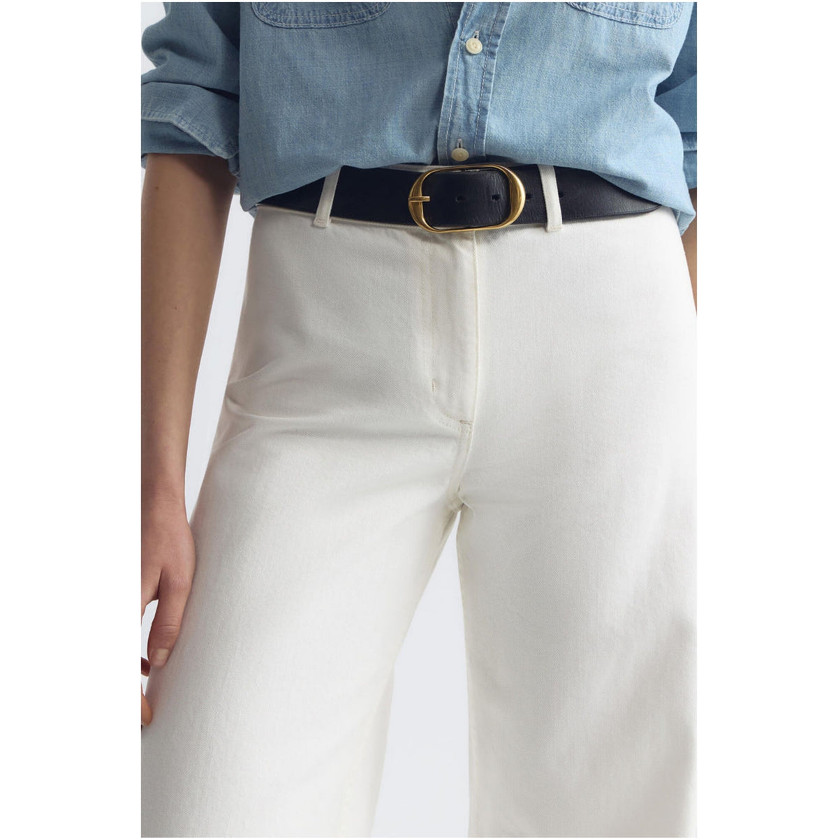 A close-up of a person wearing off-white wide-leg trousers, a black leather belt with a gold-tone oval buckle, and a light blue denim shirt.