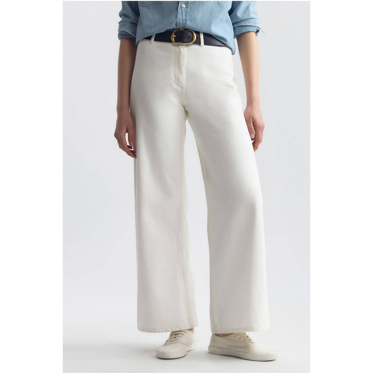 A model wearing a pair of off-white wide-leg trousers, styled with a light blue denim shirt, a black belt with a gold buckle, and off-white sneakers against a light gray studio background.