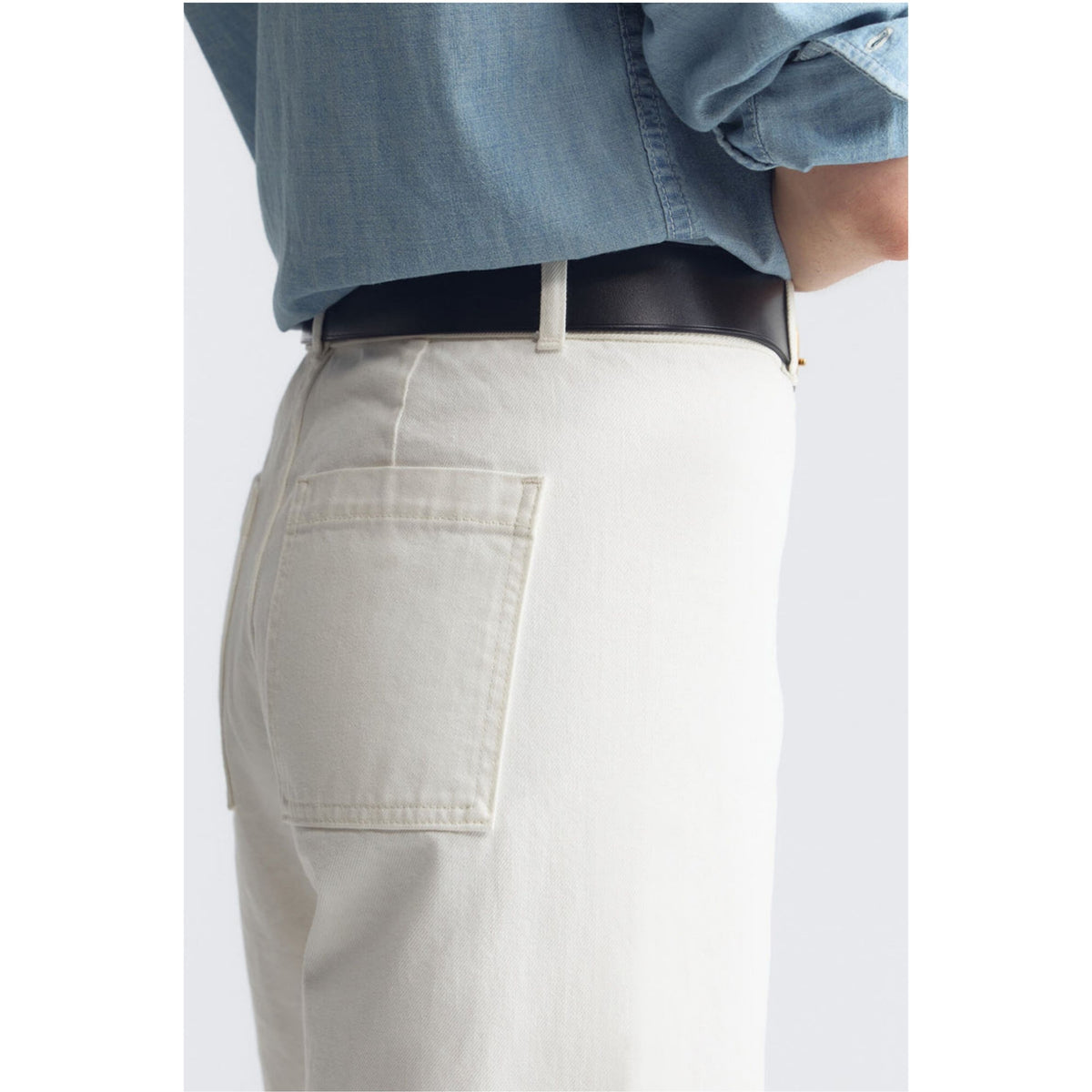 Close-up of a person wearing off-white or cream-colored trousers with a black belt and a light blue denim shirt, shown from the back.