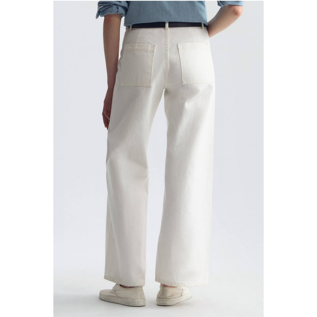 A person shown from the back wearing off-white wide-leg jeans with two large back pockets, a dark belt, a light blue denim shirt, and off-white slip-on shoes against a plain light gray studio background.
