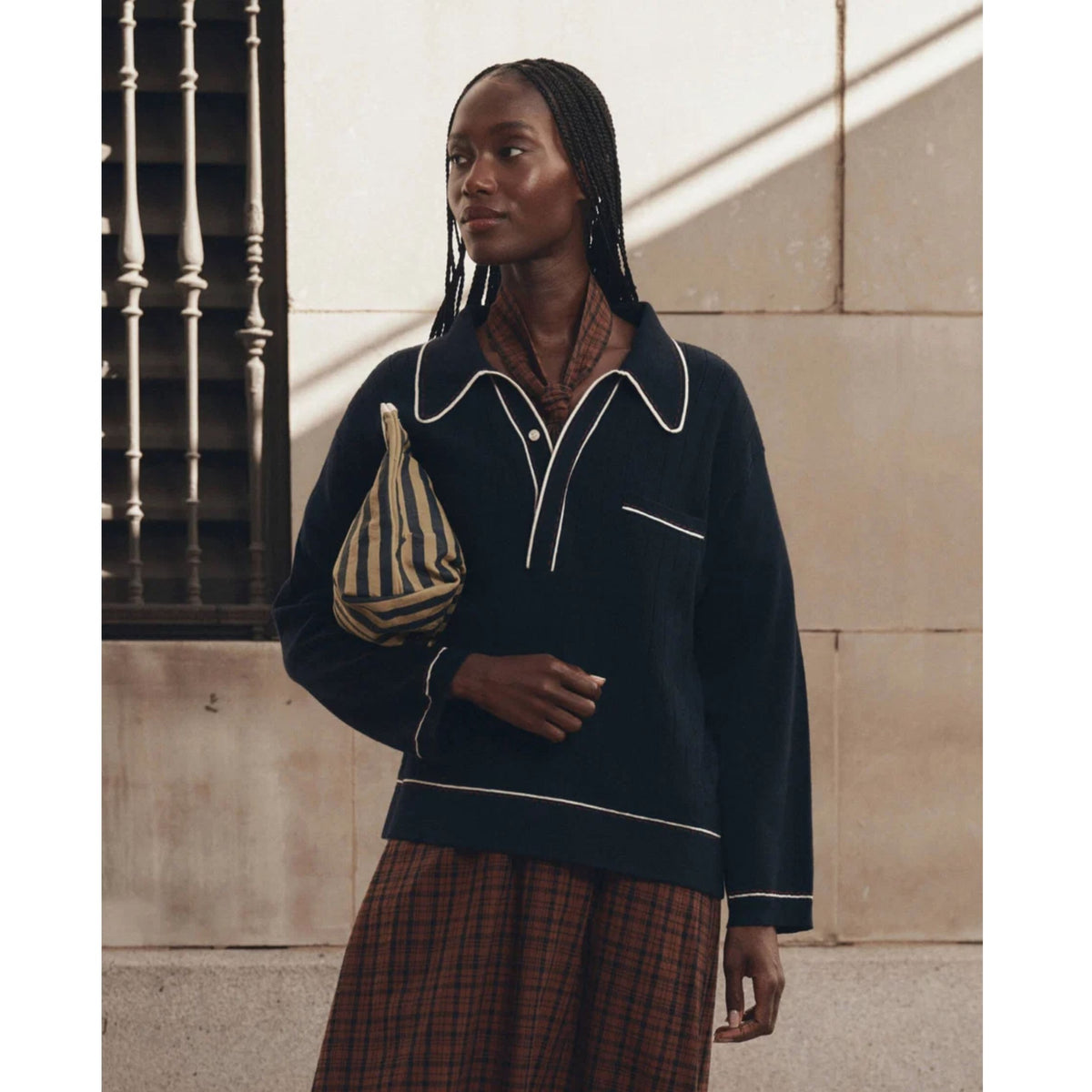 A woman stands outdoors against a beige stone wall, wearing a dark navy long-sleeve knit top with white piping and a brown plaid skirt, while carrying a beige and navy striped bag.