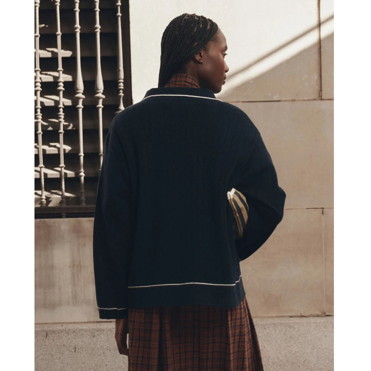 A model shown from behind wearing a dark navy blue knit cardigan with white contrast trim, layered over a brown and red plaid shirt and skirt, holding a striped clutch against a beige stone wall.