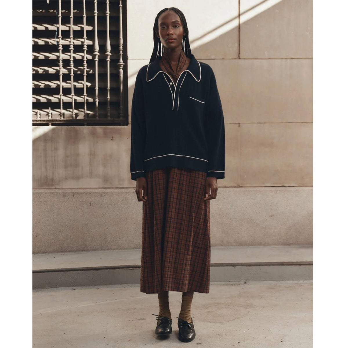 A model stands outdoors wearing a dark navy long-sleeve knit top with white piping details and a brown and red plaid midi skirt, paired with black leather shoes.