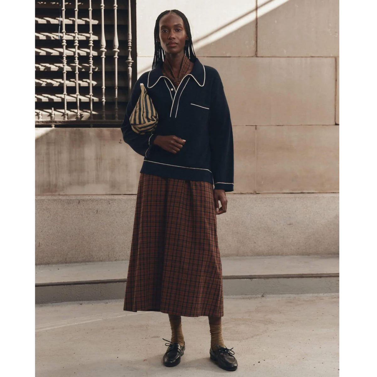 A model stands outdoors wearing a dark navy long-sleeve knit top with white piping and a brown and red plaid midi skirt, paired with brown socks and black loafers.