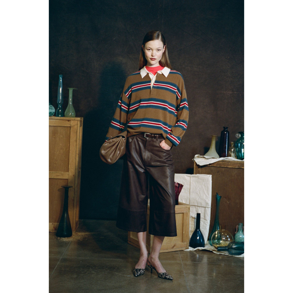 Woman wearing a striped brown sweater with a white collar and brown leather long shorts, holding a brown back standing in a room with boxes and vases.