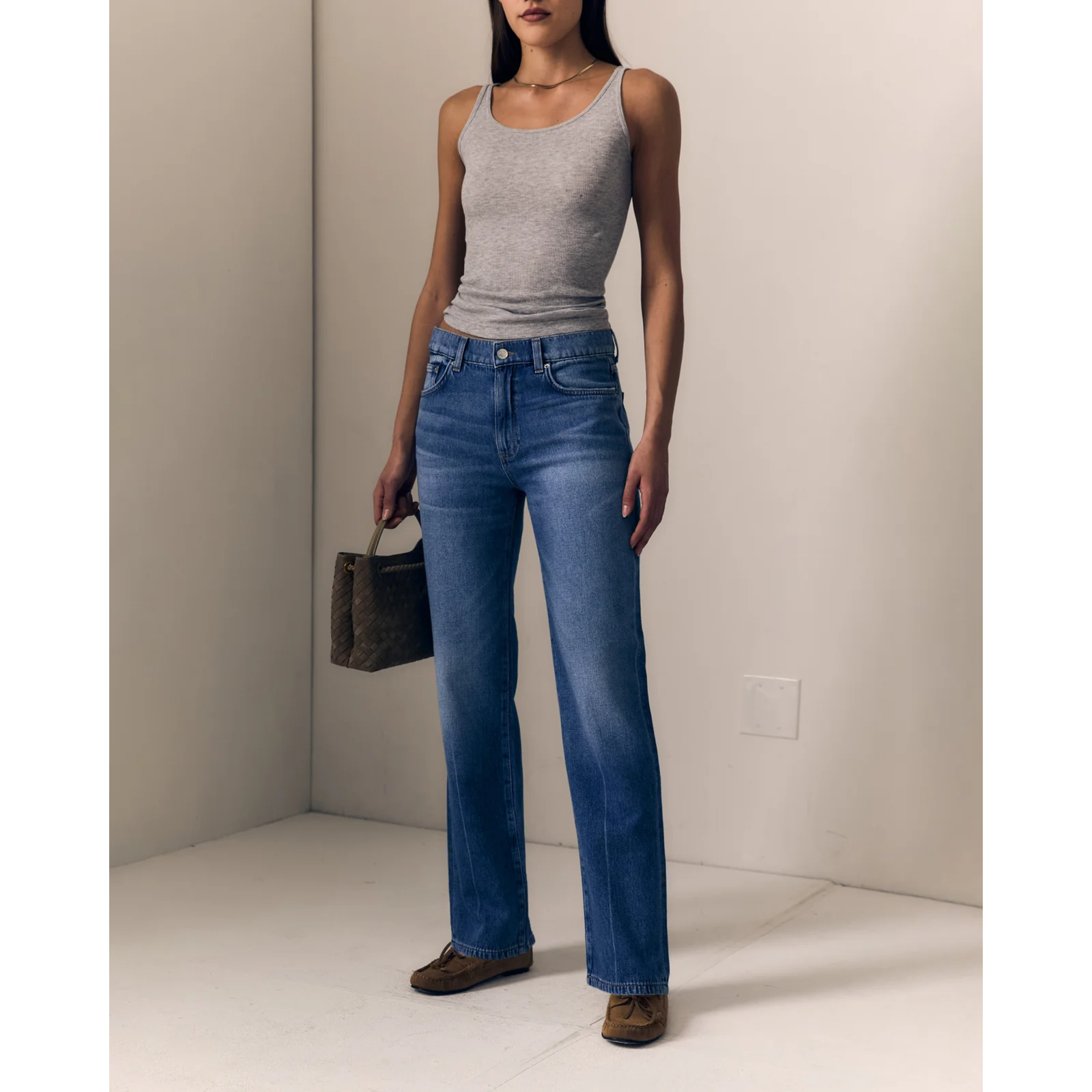 A woman modeling a pair of medium-wash blue straight-leg jeans with a gray ribbed tank top and brown suede shoes, holding a dark patterned handbag in a minimalist beige room.