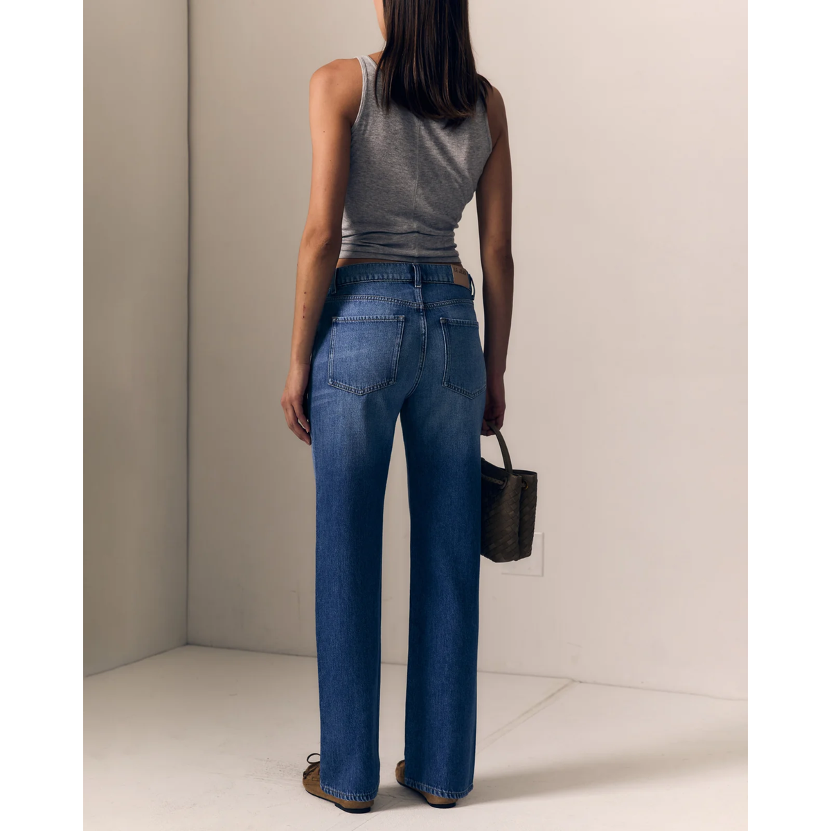 A woman modeling a pair of medium-wash blue straight-leg jeans with a gray ribbed tank top and brown suede shoes, holding a dark patterned handbag in a minimalist beige room.